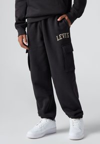 Černé tepláky s volným střihem, se zlat�ým logem LEVI'S, bočními cargo kapsami a elastickými manžetami. V páru s bílými teniskami.