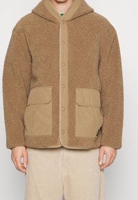 Chaqueta de forro polar marrón con exterior texturizado, forro beige y dos bolsillos frontales. Presenta una capucha y cierre con botones a presión.