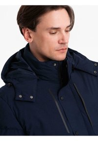 Muž v tmavě modré zimní bundě s kapucí, na zapínání na knoflíky a zip, dívající se dolů na bílém pozadí.