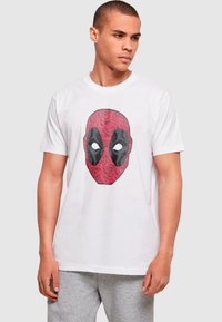 Witte katoenen t-shirt met een rood en zwart grafisch ontwerp van een gemaskerd gezicht met opvallende ogen, tegen een achtergrond van bloemenpatroon.