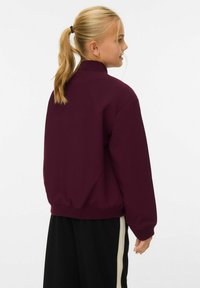 Felpa bordeaux con colletto alto, polsini e orlo a costine, realizzata in tessuto morbido, indossata sopra pantaloni neri con strisce laterali.