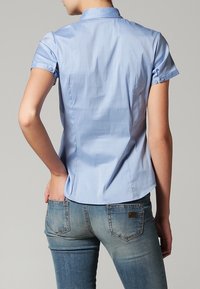Chemise bleu clair à manches courtes avec col, coupe ajustée, détails de poignets boutonnés et texture lisse. Portée avec un jean bleu délavé.