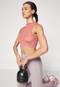Ανοιχτό ροζ αθλητικό crop top με λογότυπο, συνδυασμένο με ανοιχτό μωβ κολάν. Μαύρο kettlebell κρατημένο και με τα δύο χέρια, με φόντο μίνιμαλ στούντιο.