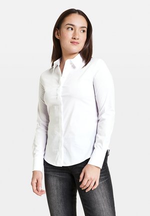 Weißes Langarmhemd aus Baumwolle mit Button-Down-Kragen, abgerundetem Saum und dezenten Ärmeldetails. Trägt dazu eine dunkle Jeans.