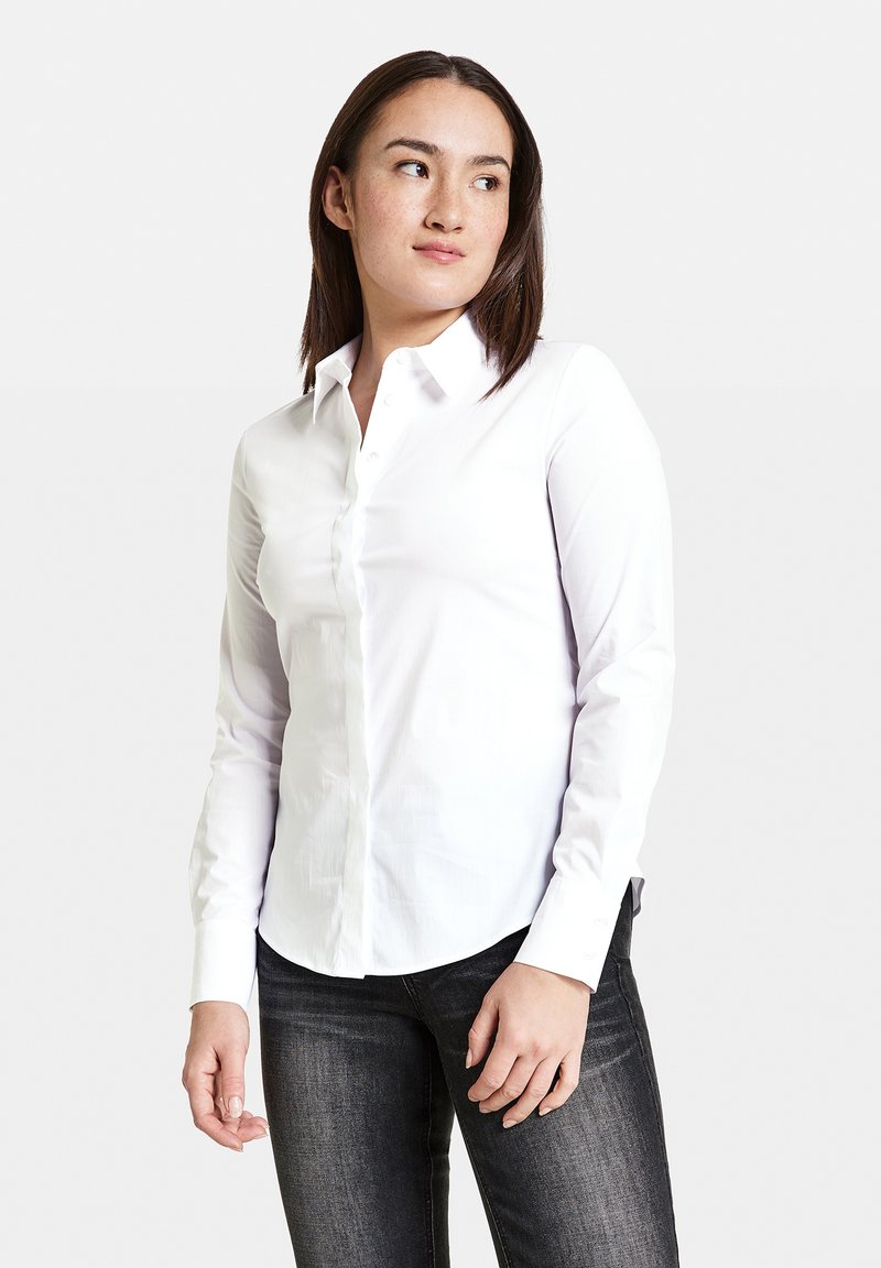 Weißes Langarmhemd aus Baumwolle mit Button-Down-Kragen, abgerundetem Saum und dezenten Ärmeldetails. Trägt dazu eine dunkle Jeans.