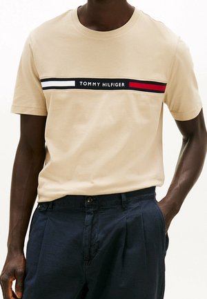 Beige cotton t-shirt with a crew neck, featuring a horizontal navy, white, and red striped graphic across the chest with "TOMMY HILFIGER" text.