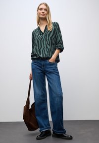 Blusa a righe verde e nera con scollo a V, abbinata a jeans blu a gamba larga. Una borsa a tracolla marrone e scarpe nere completano l'outfit.
