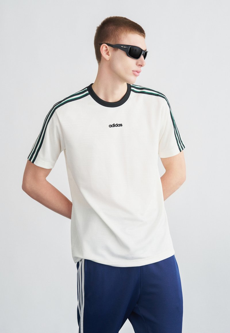 Valge lühikeste varrukatega T-särk, musta kraega ja kolme rohelise triibuga kummalgi õlal. Must "adidas" logo rinnal. Tumedasinised püksid.