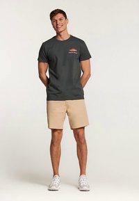 Dunkelgraues T-Shirt mit einem roten Grafiklogo mit der Aufschrift "CATCH OF THE DAY" auf der linken Brust. Paar beige Shorts und weiße Turnschuhe.