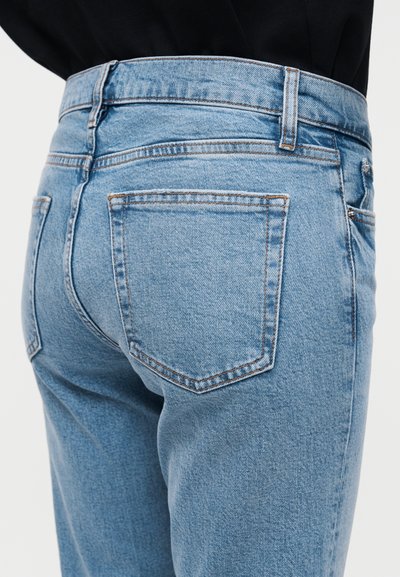 Gros plan sur un jean en denim bleu porté avec un haut noir, montrant la poche arrière et les passants de ceinture sur un fond blanc uni.