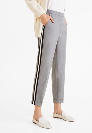 Kobieta nosząca szare krótkie spodnie z czarno-białymi bocznymi paskami, białe buty wsuwane, biały top i beżowy kardigan, ręce w kieszeniach.