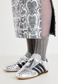 Hopeiset metalliset urheilukengät, joissa on mustat raidat, yhdistettyinä harmaisiin raidallisiin sukkiin. Kengissä on pyöreä kärki ja virtaviivainen muotoilu.