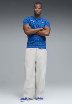 Mann mit Brille, der ein blau gemustertes Puma-T-Shirt, lockere weiße Hosen und blaue Turnschuhe trägt, steht mit verschränkten Armen.