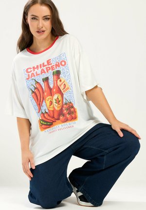 Femme agenouillée, portant un t-shirt blanc surdimensionné avec un col rouge et un graphisme de piments jalapeños, bouteilles de sauce piquante et tomates.