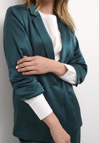 Teal satin blazer med avslappnad passform, framfickor och uppvikta ärmar över en vit skjorta med knappar. Slät yta, enkel design.