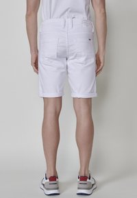Pantalones cortos de mezclilla blanca con puños enrollados, que cuentan con dos bolsillos traseros y una etiqueta en la cinturilla, mostrados desde una vista trasera.