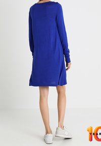 Eine Person in einem blauen Langarmkleid und weißen Sneakers steht mit dem Rücken zu einer schlichten weißen Wand.