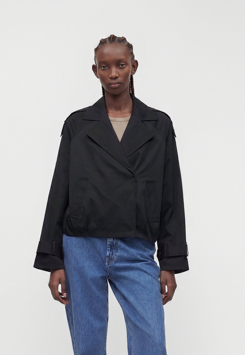 Femme aux cheveux tressés portant une veste noire oversize par-dessus un haut beige et un jean bleu, debout devant un fond clair uni.