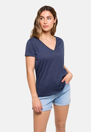 Mujer con cabello ondulado que lleva una camiseta de manga corta azul marino con cuello en V y shorts vaqueros azul claro remangados, posando con una mano en el bolsillo.