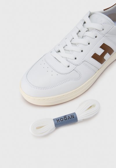 Λευκό αθλητικό παπούτσι από δέρμα με διάτρητη μύτη, καφέ λογότυπο στο πλάι και λευκά κορδόνια. Περιλαμβάνει επιπλέον λευκά κορδόνια με θήκη με λογότυπο.