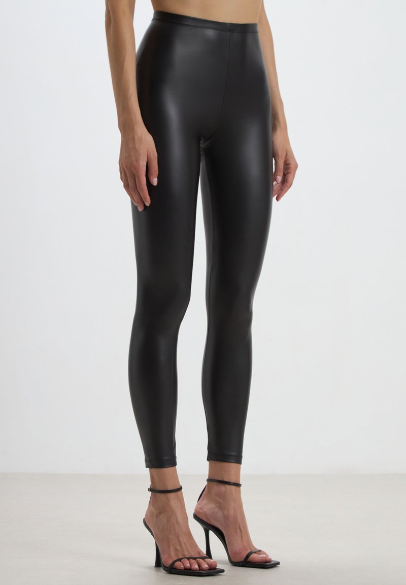 Leggings noirs taille haute fabriqués en matériau brillant et extensible, moulants sur les jambes, assortis à des sandales à talons hauts noires à lanières.