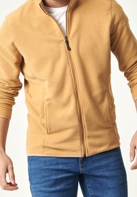 Giacca in pile beige, con zip integrale e colletto alto, due tasche laterali e polsini a costine, caratterizzata da una texture liscia.