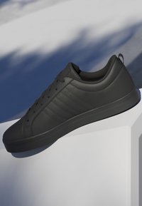 Baskets noires basses avec design à lacets, col rembourré et détails texturés, positionnées sur une surface blanche avec un arrière-plan d'ombre bleue.