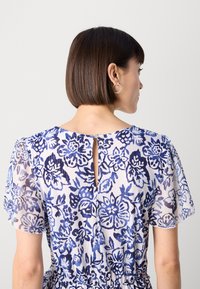 Robe florale en tissu transparent avec des motifs bleus et blancs, détail du dos en forme de clé, manches courtes et design taille froncée.