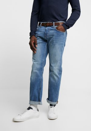 Mand iført lyseblå opkørte jeans, hvide sneakers, hvide sokker, et mørkebrunt bælte og en mørk langærmet skjorte med den ene hånd i lommen.