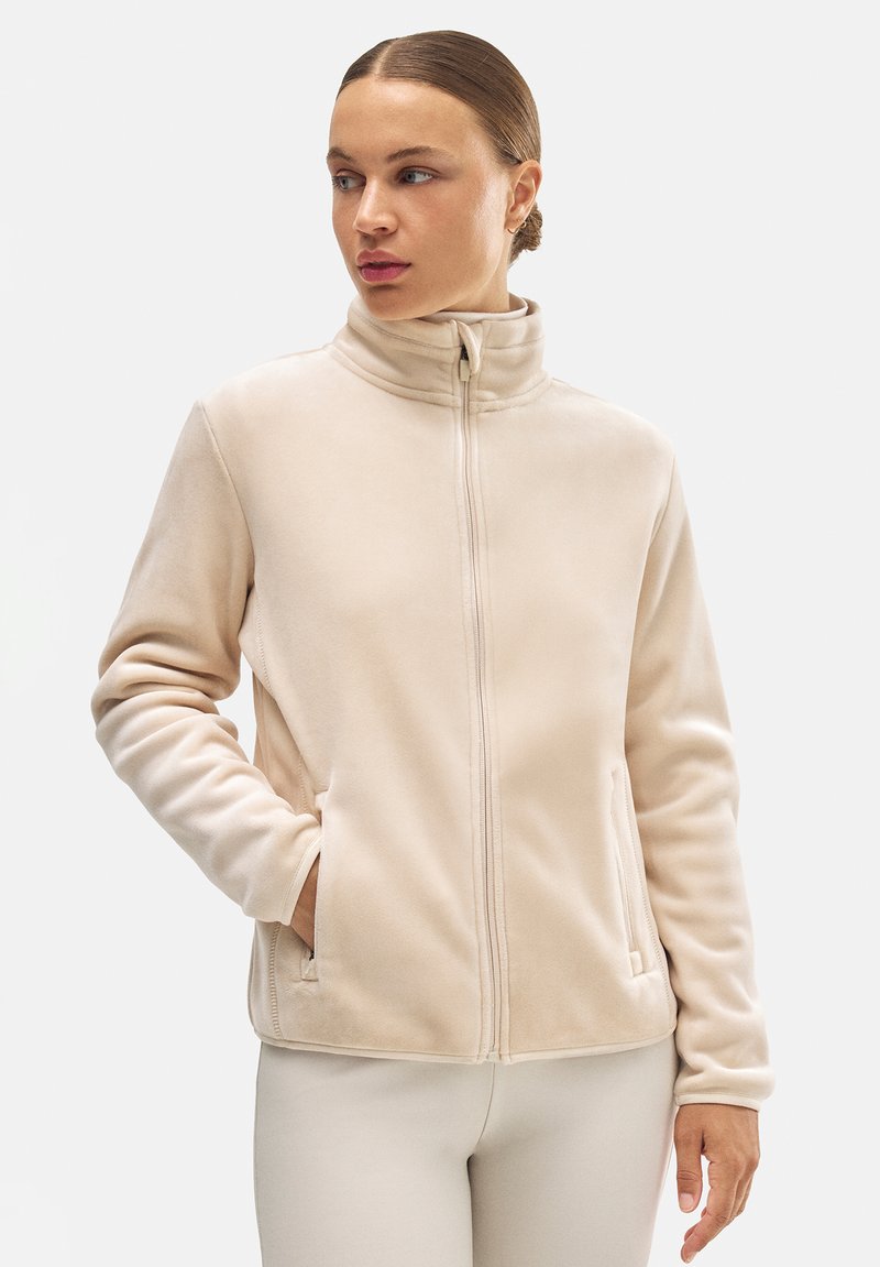 Chaqueta de forro polar beige con cuello alto, cierre de cremallera y bolsillos laterales; textura suave; mangas largas; silueta ajustada.