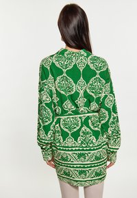 Žalias raštuotas tunikas su ilgomis rankovėmis, puoštas gėlių raštu kreminės spalvos ir su juosmens juostele. Medžiaga yra glotni.