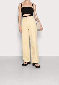 Zwart geribbelde crop top met dunne bandjes, gecombineerd met gele corduroy broek met hoge taille en wijde pijpen. Model houdt een beige tas vast en draagt zwarte slippers.
