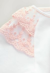 Biała bluzka z delikatnym różowym haftowanym koronkowym zdobieniem na jednym ramieniu, o gładkiej fakturze i prostym, okrągłym dekolcie.