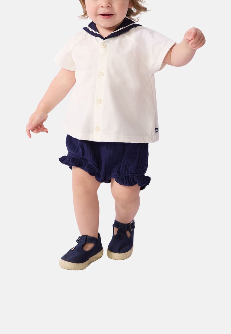 Camicia bianca a maniche corte con colletto blu navy, abbinata a pantaloncini blu scuro con volant e scarpe blu navy con dettagli ritagliati e suole chiare.