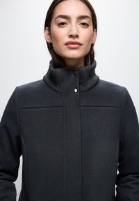 Schwarzer Mantel mit hohem Kragen, strukturiertem Stoff und Knopfverschluss. Besitzt maßgeschneiderte Nähte und eine glatte Oberfläche.