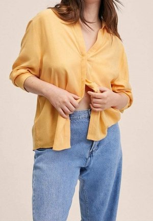 Person iført løs gul skjorte med knapper og lyseblå jeans med høj talje, der holder skjorten let åben ved taljen.