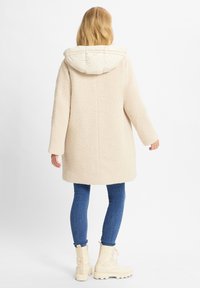 Cappotto beige e morbido con cappuccio a fantasia crema, maniche lunghe e taglio dritto, abbinato a jeans blu e stivaletti beige con lacci.