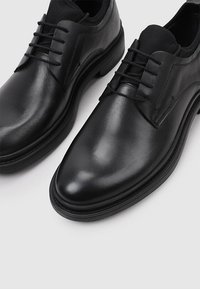 Chaussures en cuir noir avec une texture lisse, bout pointu et quatre œillets pour lacets. Équipées d'une semelle en caoutchouc et de coutures minimales.