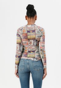 Long-sleeve mesh top featuring a collage of vintage gas station advertisements in various colors. Paired with blue denim jeans.