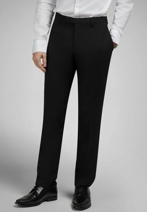 Mann in schwarzen Anzughosen, schwarzen Lederschuhen und weißem, langärmligem Hemd mit Knöpfen, steht mit den Händen nahe den Taschen.