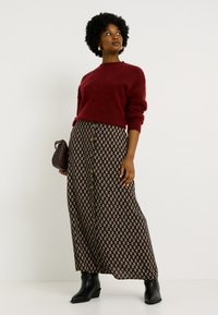 Pull en tricot bourgogne à col rond assorti à une jupe maxi noire à motifs, ornée de boutons dorés et de motifs floraux. Bottines noires