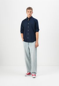 Joven de pie con las manos en los bolsillos, vistiendo una camisa de botones azul marino, jeans de color azul claro y zapatillas rojas, contra un fondo blanco.
