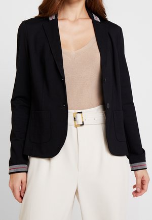 Frau trägt einen schwarzen Blazer mit gestreiften Manschetten über einem beigen Stricktop, kombiniert mit hoch taillierten weißen Hosen mit breitem Gürtel und quadratischer Schnalle.