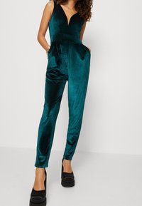 Teal sammetsoverall med V-ringning, figursydd midja, avsmalnande ben och sidofickor. Stylerad med svarta plattformsskor.