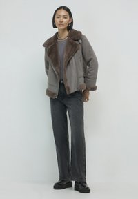 Giacca in shearling marrone con ampio colletto, chiusura con zip e rifiniture in pelliccia, abbinata a jeans neri a vita alta e scarpe con zeppa nere.