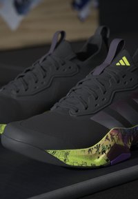 Paire de chaussures de sport noires avec des accents violets et des semelles intermédiaires à motifs jaune-vert sur une surface sombre.