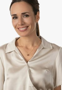 Satinbluse in Hellbeige mit Schalkragen; zarte goldene Halskette mit einem runden braunen Anhänger. Glatte Textur und entspannter Sitz.