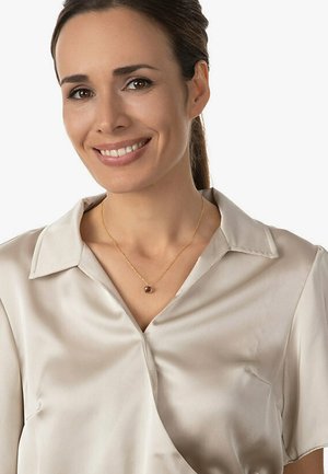 Satinbluse in Hellbeige mit Schalkragen; zarte goldene Halskette mit einem runden braunen Anhänger. Glatte Textur und entspannter Sitz.