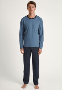 Blauwe heren pyjama top met lange mouwen en een borstzak, gecombineerd met donker marineblauwe broek. Zachte stof, comfortabele pasvorm, eenvoudig ontwerp.