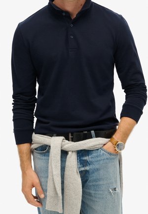 Mann trägt ein marineblaues Langarm-Poloshirt, blaue Jeans mit abgenutztem Fleck, grauen Pullover um die Taille gebunden, schwarzen Gürtel und braune Lederarmbanduhr.